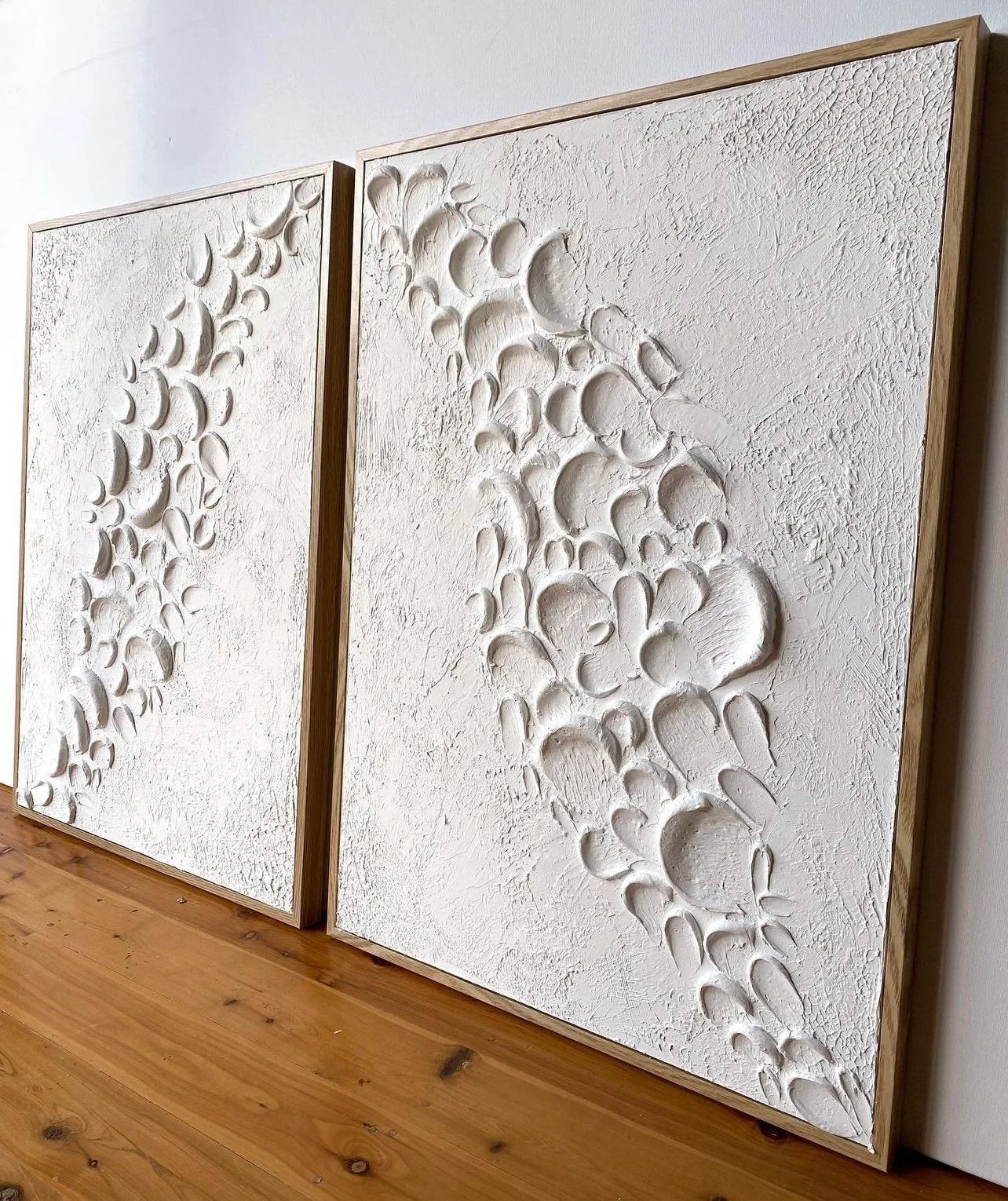 Two white textured wall artworks on a wooden surface with a white wall background.