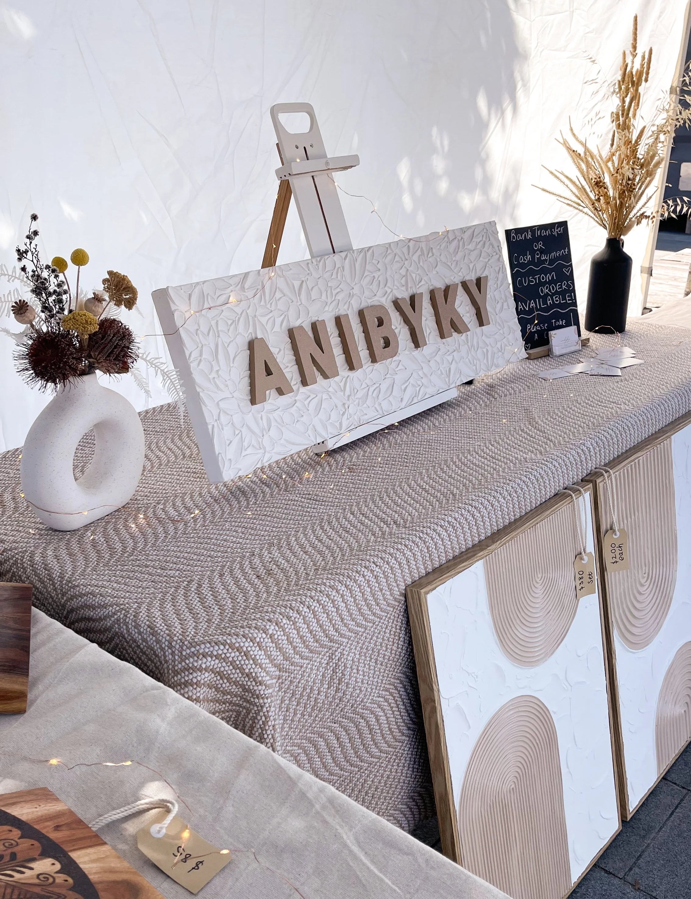 Decorative setup with 'ANIBYKY' sign, plants, and art on a market stall table.