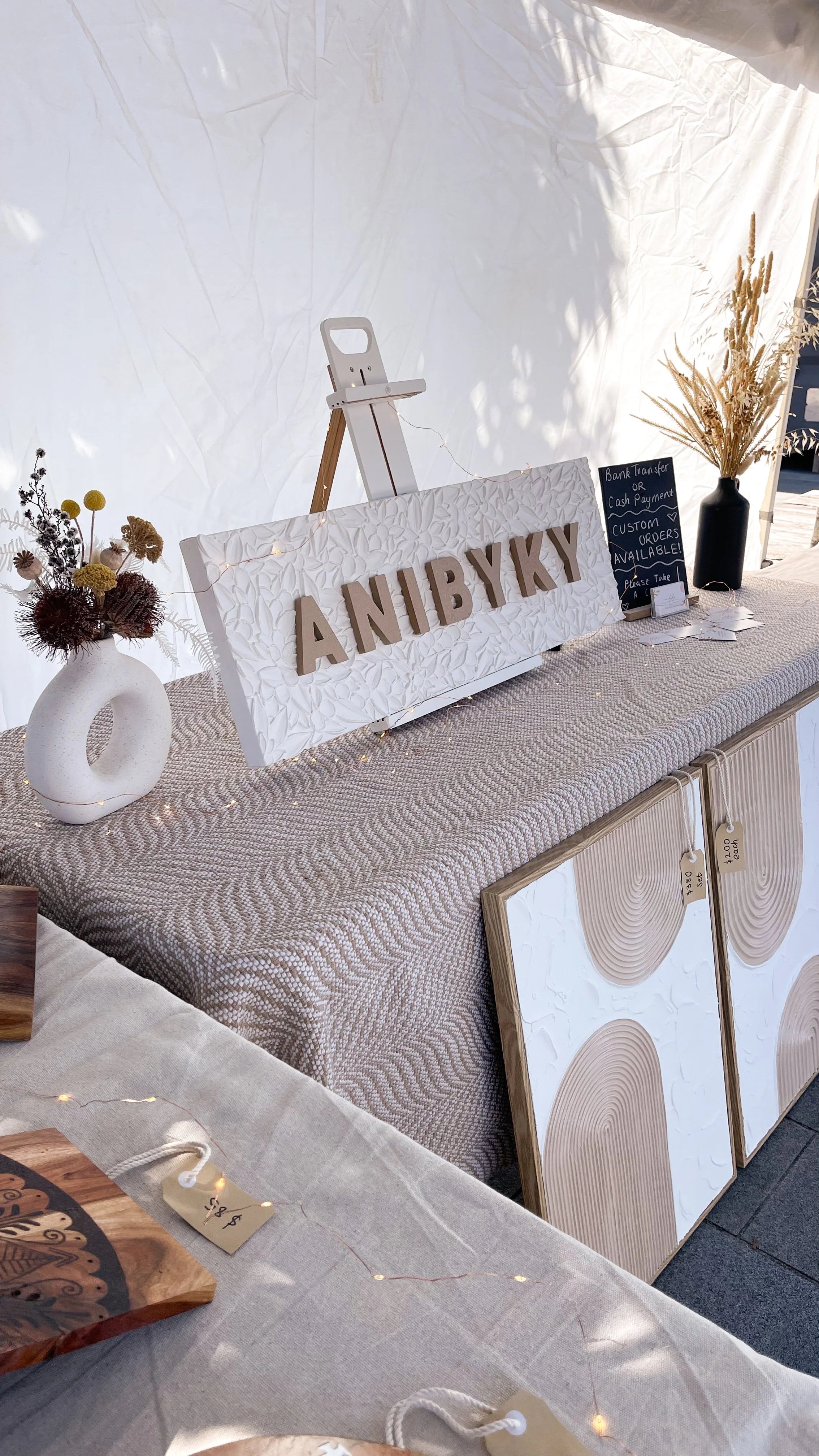 Decorative setup with 'ANIBYKY' sign, plants, and art on a market stall table.