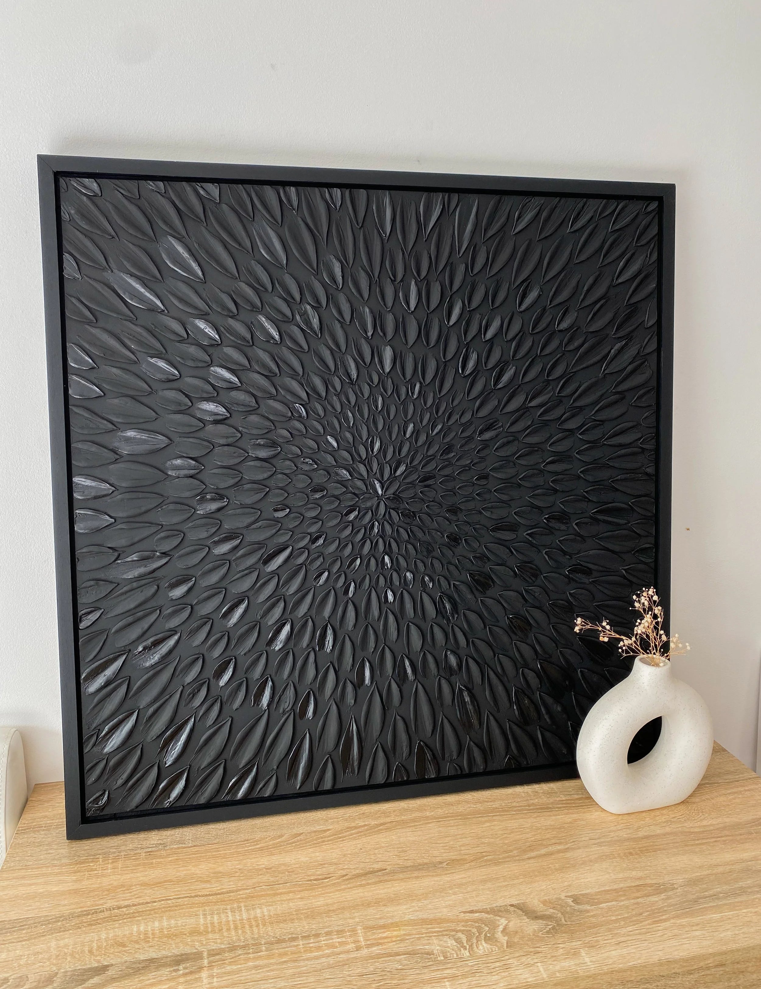 Black textured art piece on a wooden surface with a white vase.