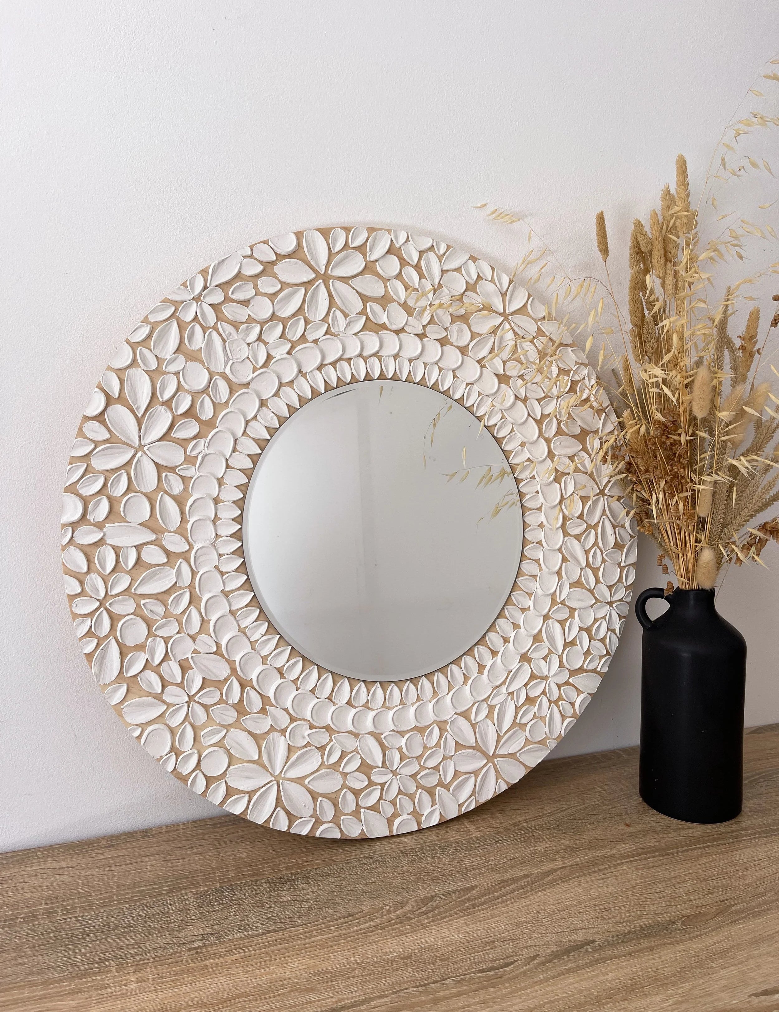 Decorative round mirror with floral textured art on a wooden surface next to a black vase with dried plants.