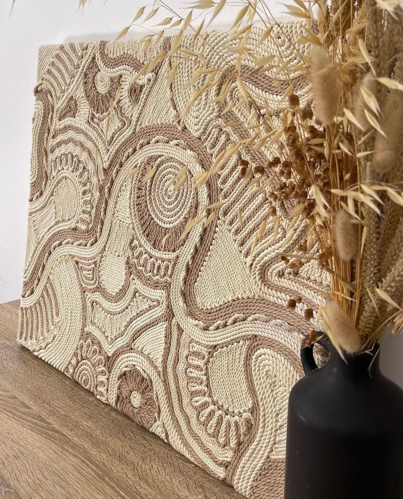 Textured wall art with intricate patterns next to a black vase with dried plants.